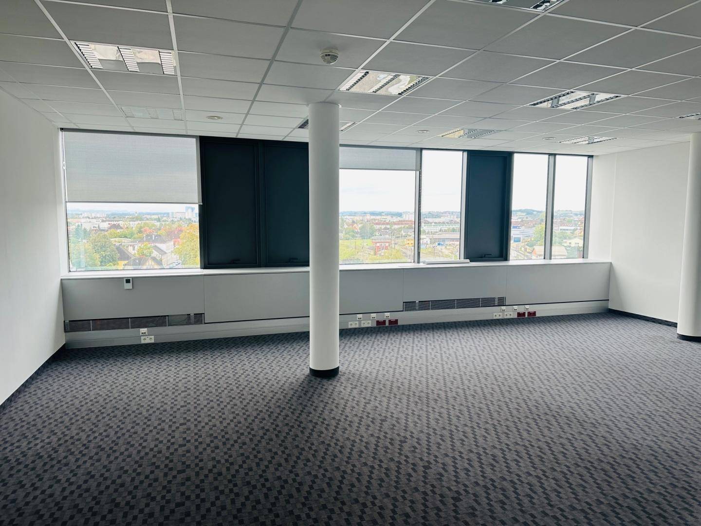 Helles Büro mit Teppichboden, Säule und breiten Fenstern mit Stadtblick.