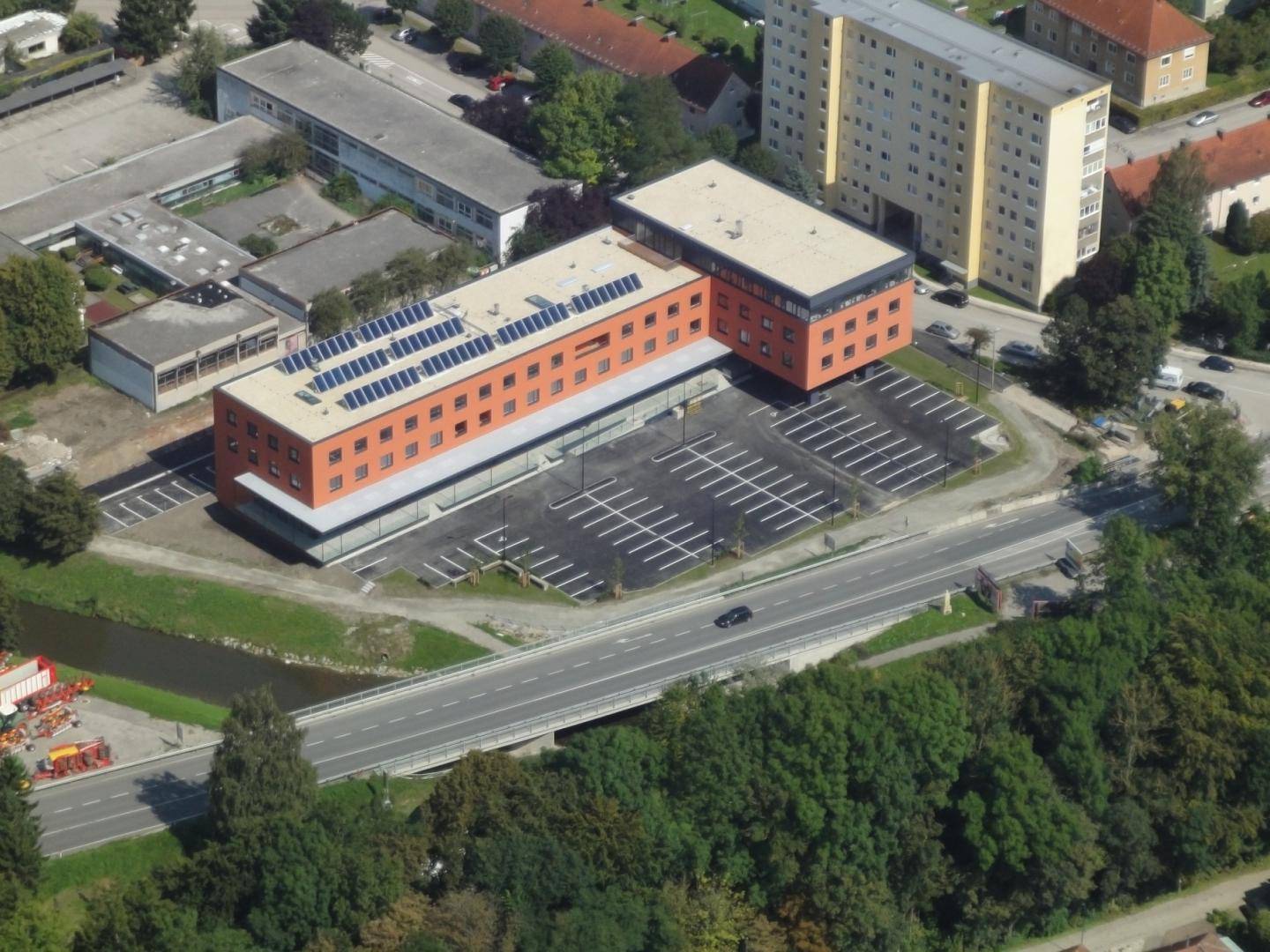 Luftaufnahme eines modernen Gebäudes mit Photovoltaikanlage auf dem Dach und großem Parkplatz.