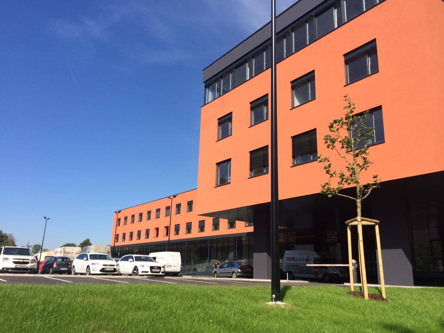 Modernes Geschäftsgebäude mit orangefarbener Fassade und Parkplätzen unter blauem Himmel.