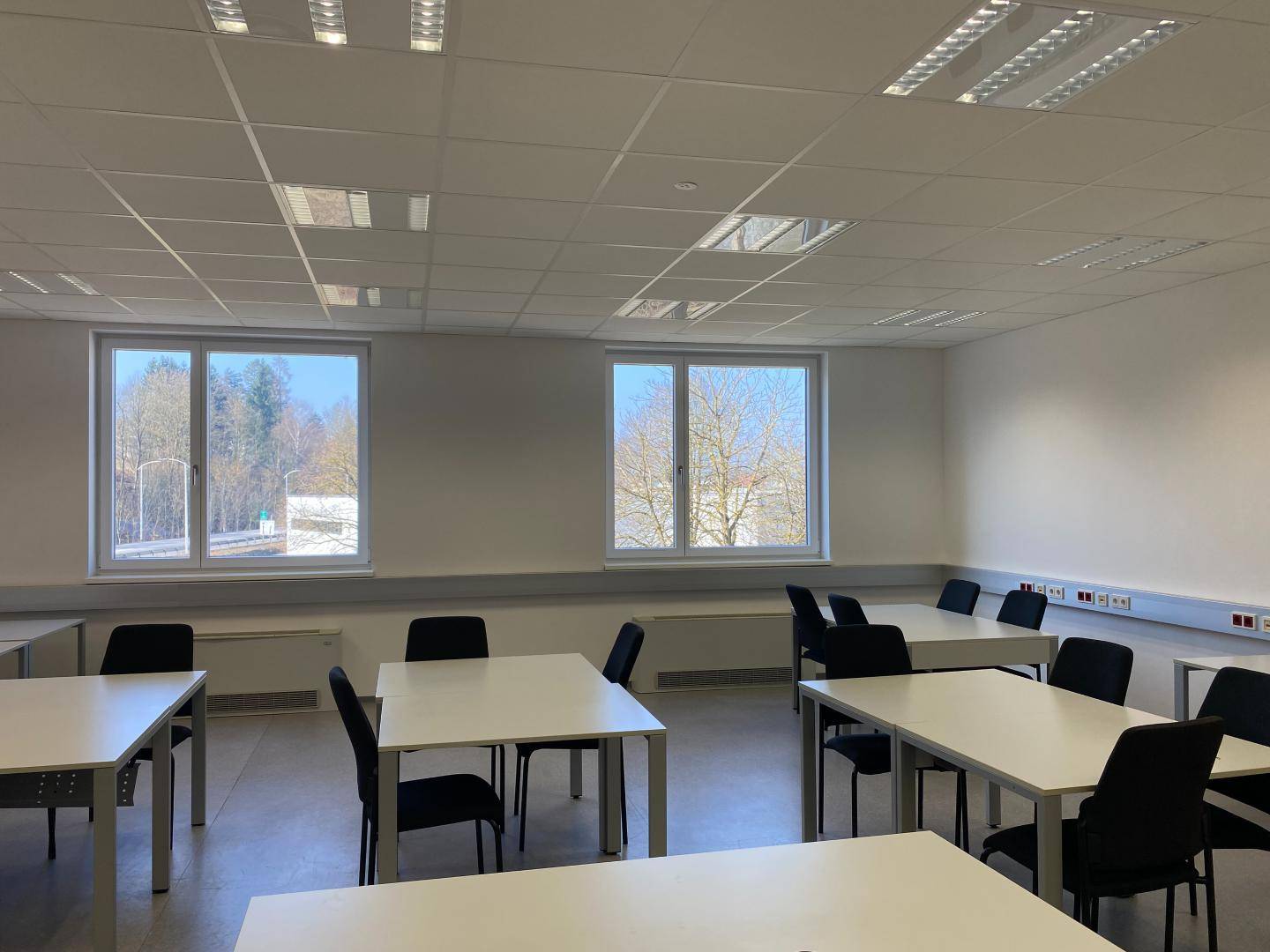 Heller Schulungsraum mit Tischen, Stühlen und großen Fenstern, die viel Tageslicht hereinlassen.