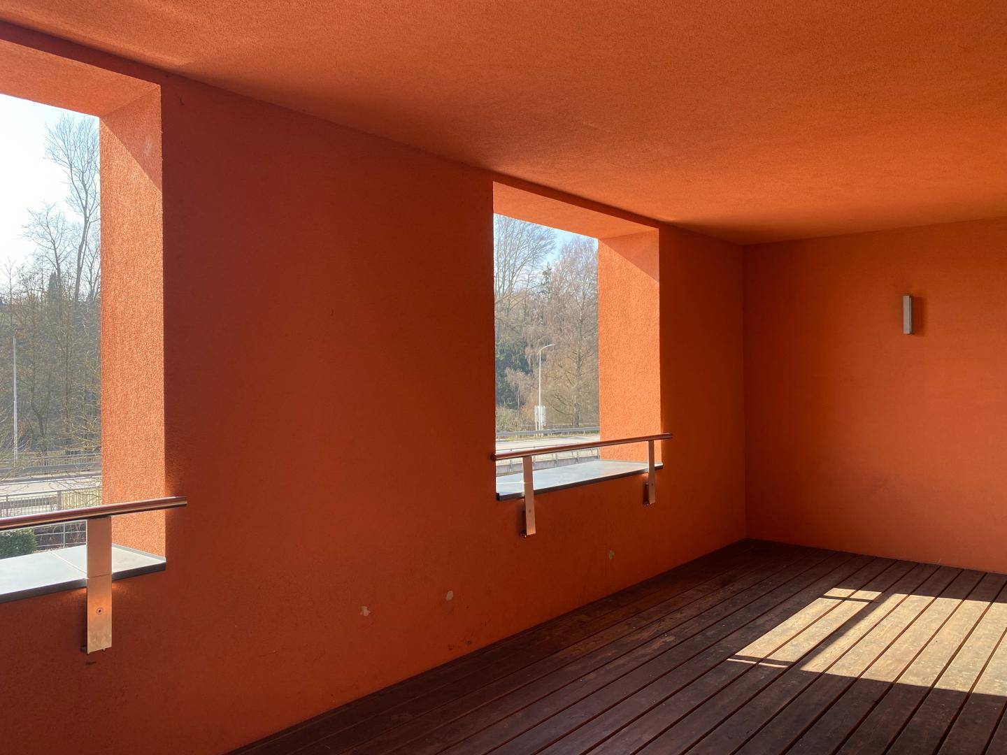 Überdachter Außenbereich mit Holzboden und orangefarbenen Wänden, bietet Blick ins Grüne.