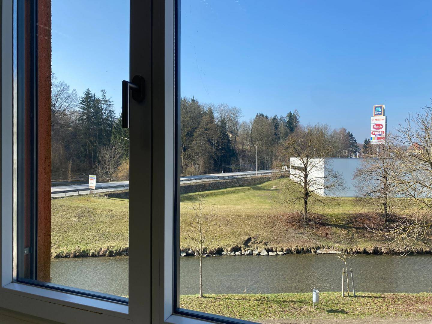 Blick aus dem Fenster auf einen Fluss, Bäume und eine Straße mit einem Supermarkt in der Ferne.