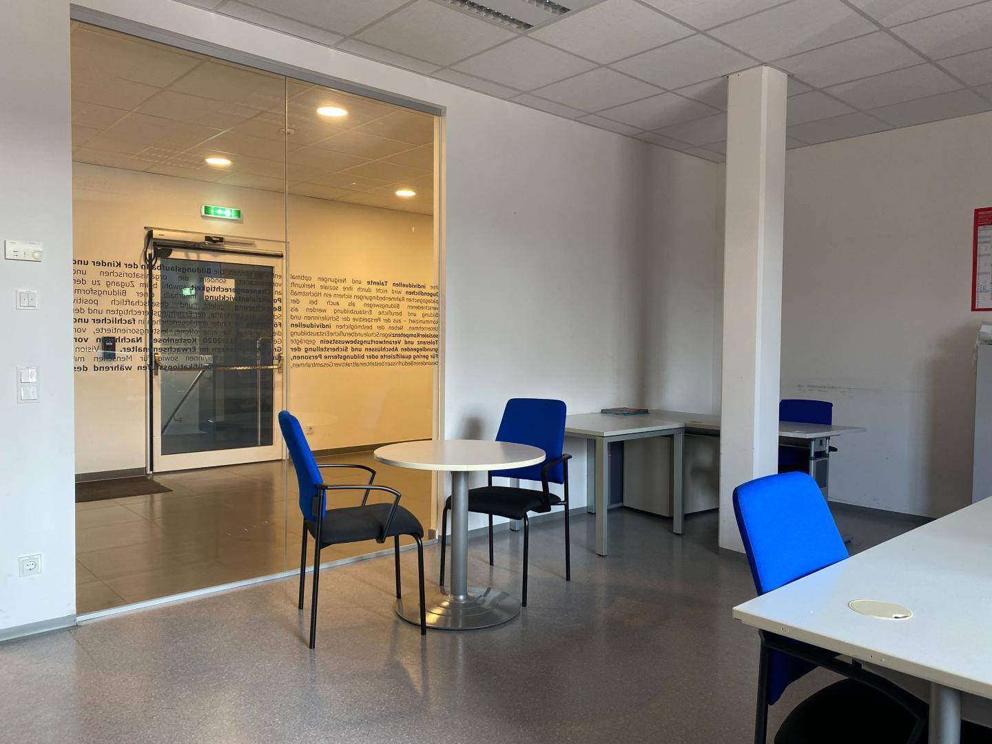 Empfangsbereich mit Glastür, kleinen Tischen und blauen Stühlen, hell und modern gestaltet.