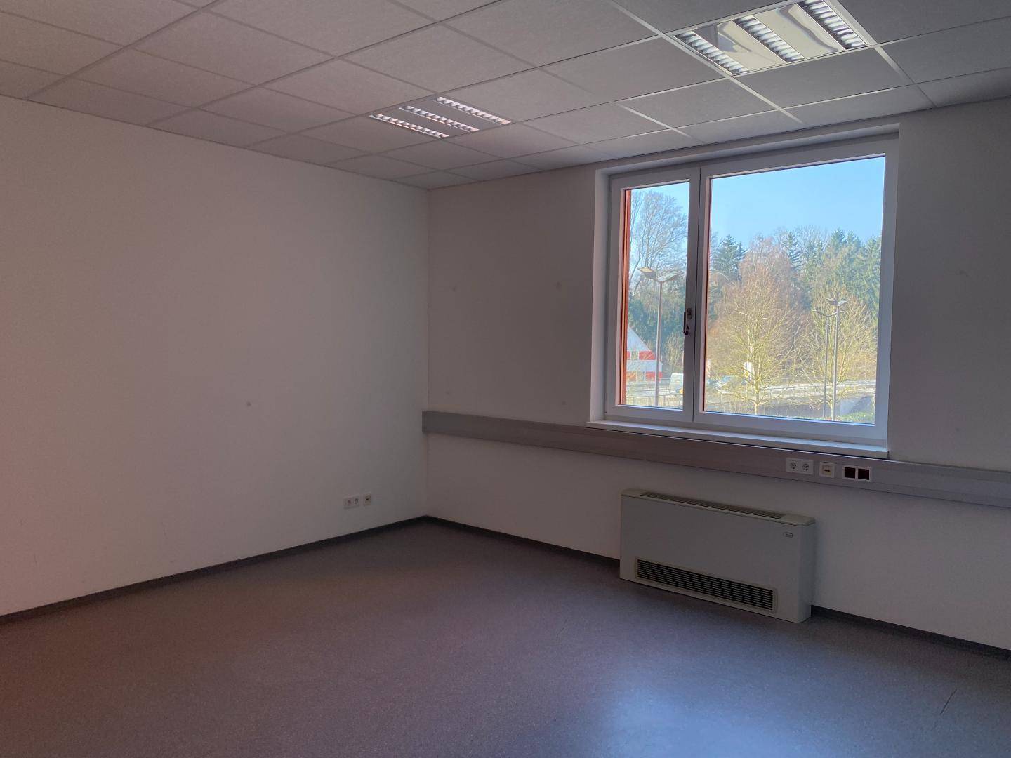 Leerer Büroraum mit einem großen Fenster, Heizkörper und grauen Bodenbelag.