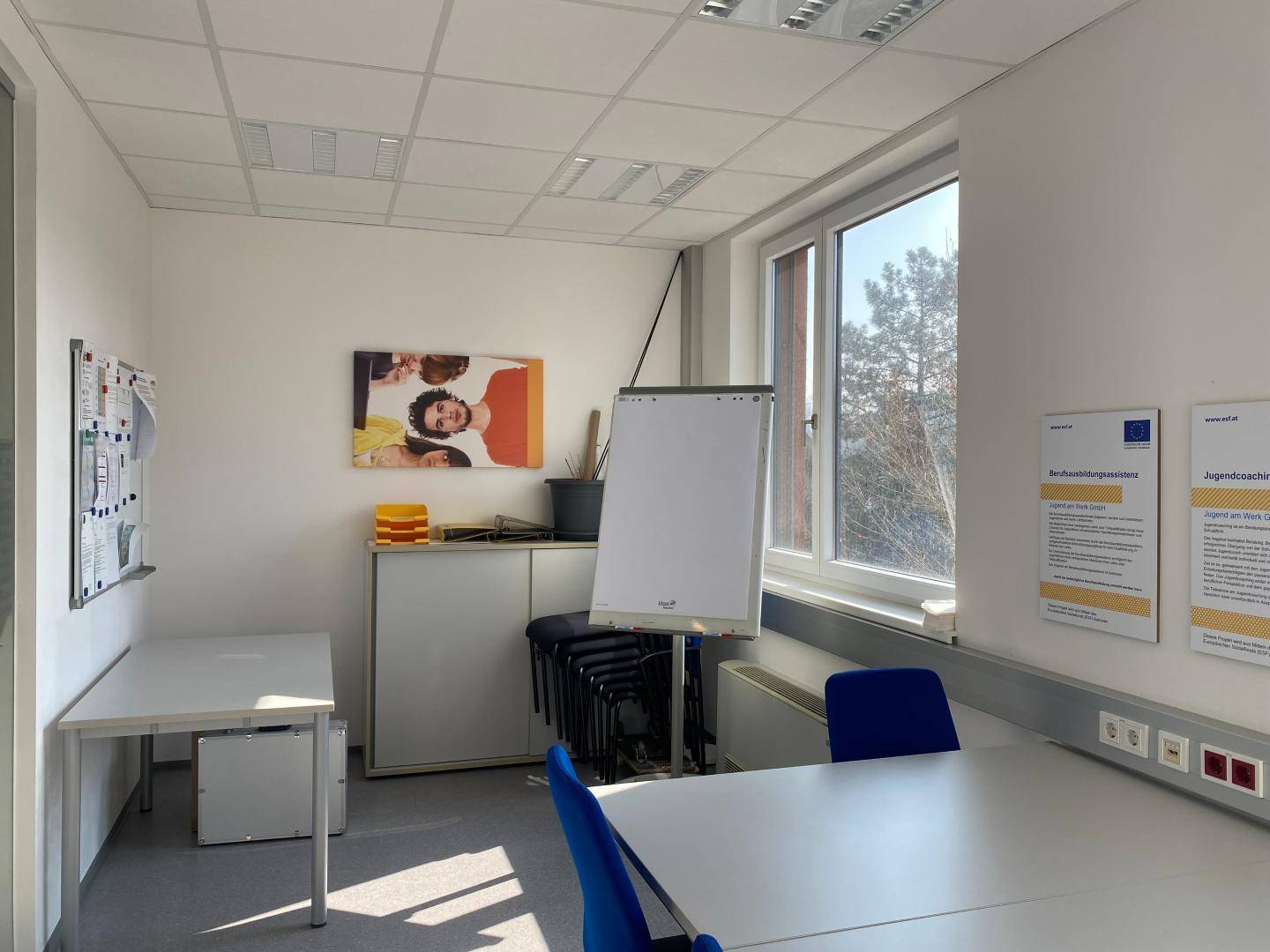 Heller Büroraum mit Schreibtischen, Stühlen, einem Whiteboard und einem großen Fenster.