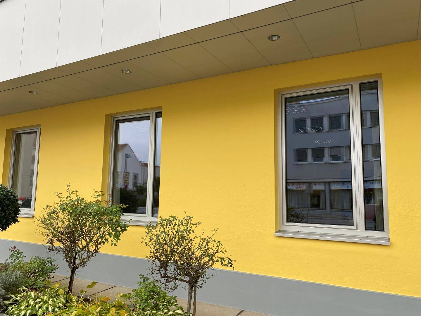 Gelbe Gebäudefassade mit modernen Fenstern und kleinen Sträuchern im Vorgartenbereich.