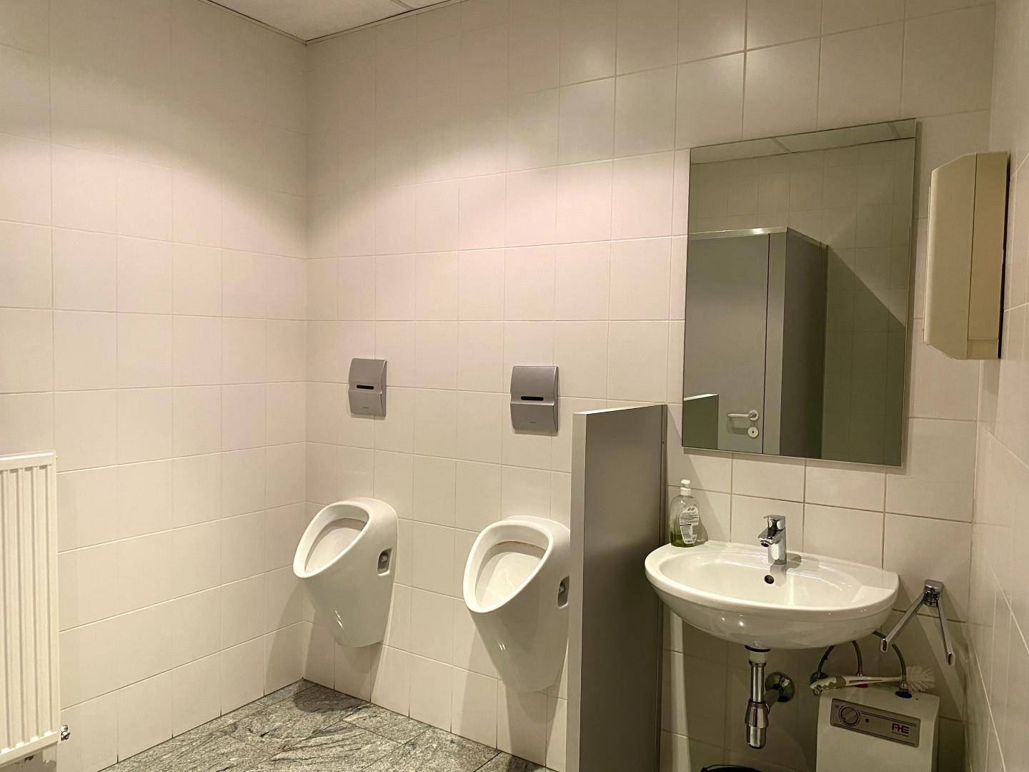 Öffentlicher Waschraum mit zwei Urinalen, Waschbecken und Spiegel.