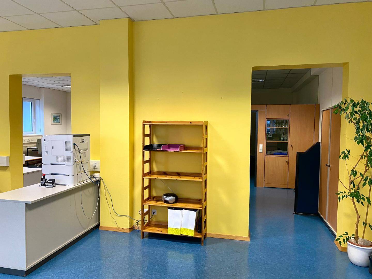Bürobereich mit gelben Wänden, Holzregal und Drucker auf einem Schrank.
