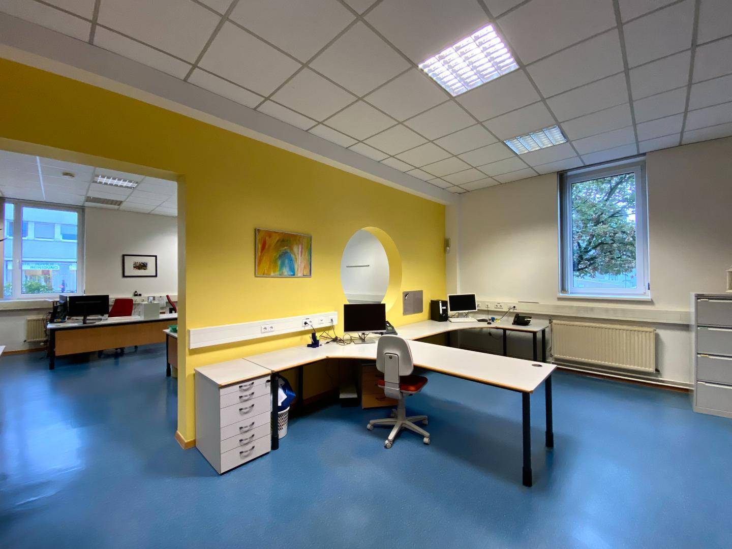 Helles Büro mit gelber Akzentwand, Eckschreibtisch und blauen Bodenbelägen.