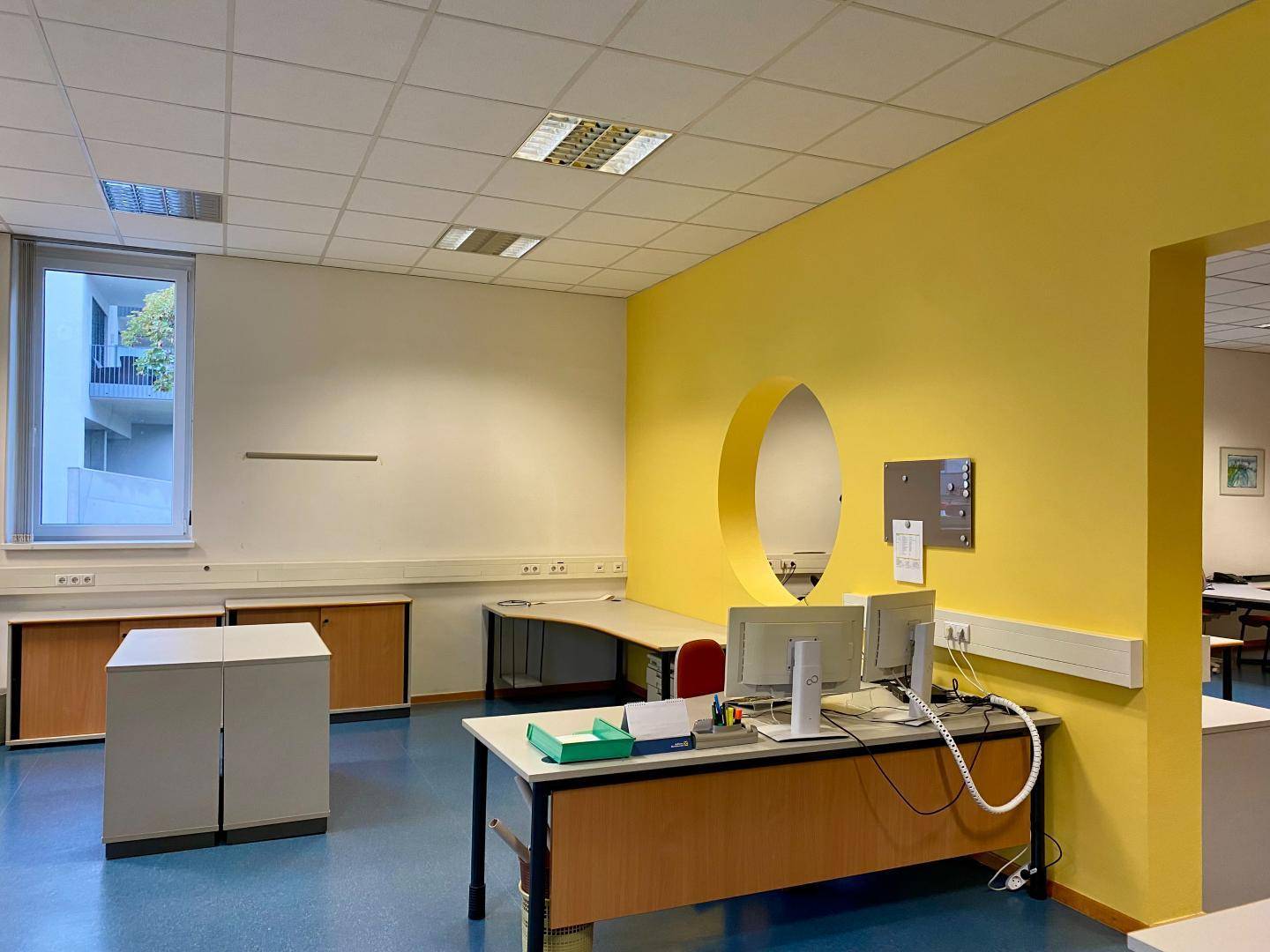 Büro mit gelber Akzentwand, runder Öffnung und mehreren Arbeitsplätzen.