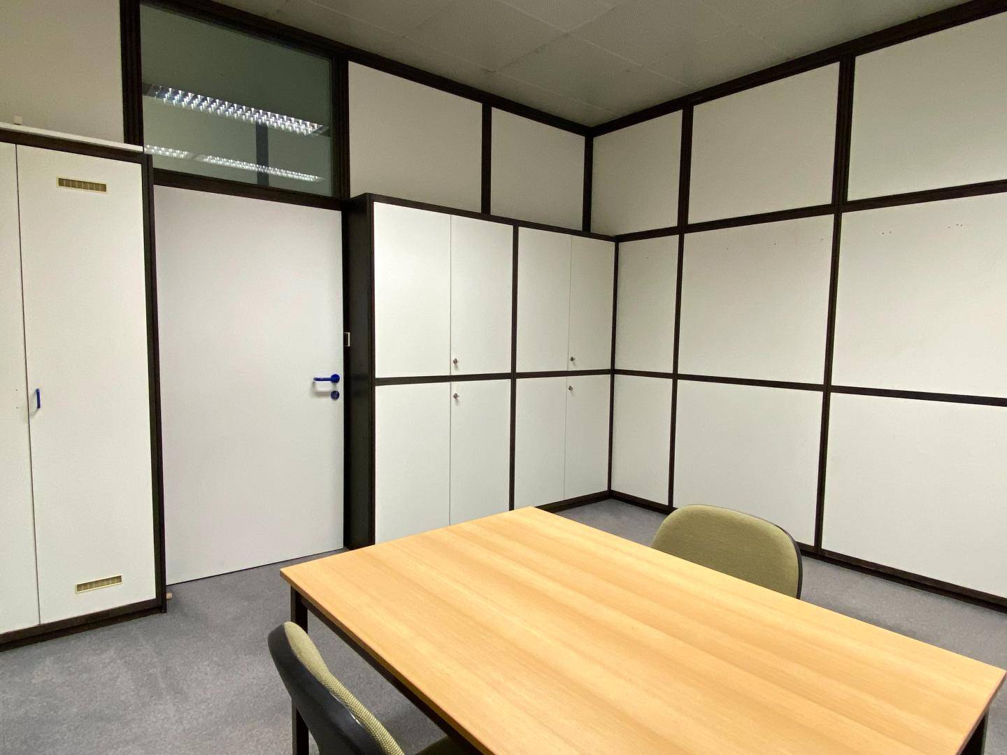 Büro mit modularen Trennwänden, integrierten Schränken und einem Holztisch in der Mitte des Raumes.