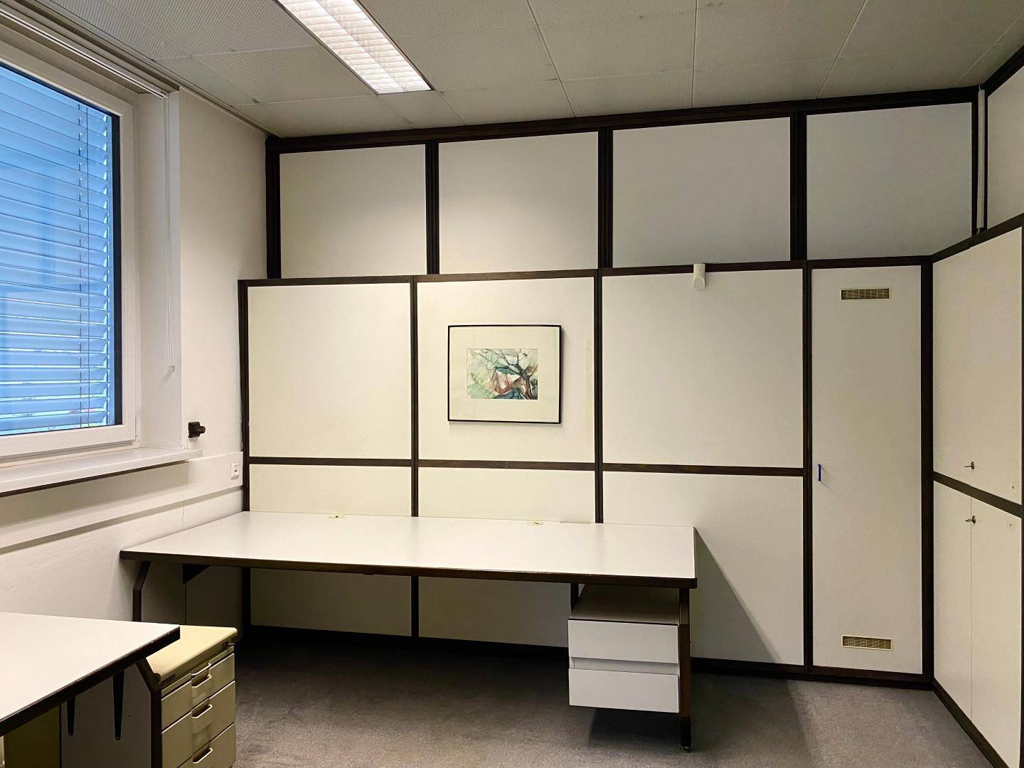 Büro mit einem Schreibtisch, integrierten Schränken und einem Fenster mit Jalousien.