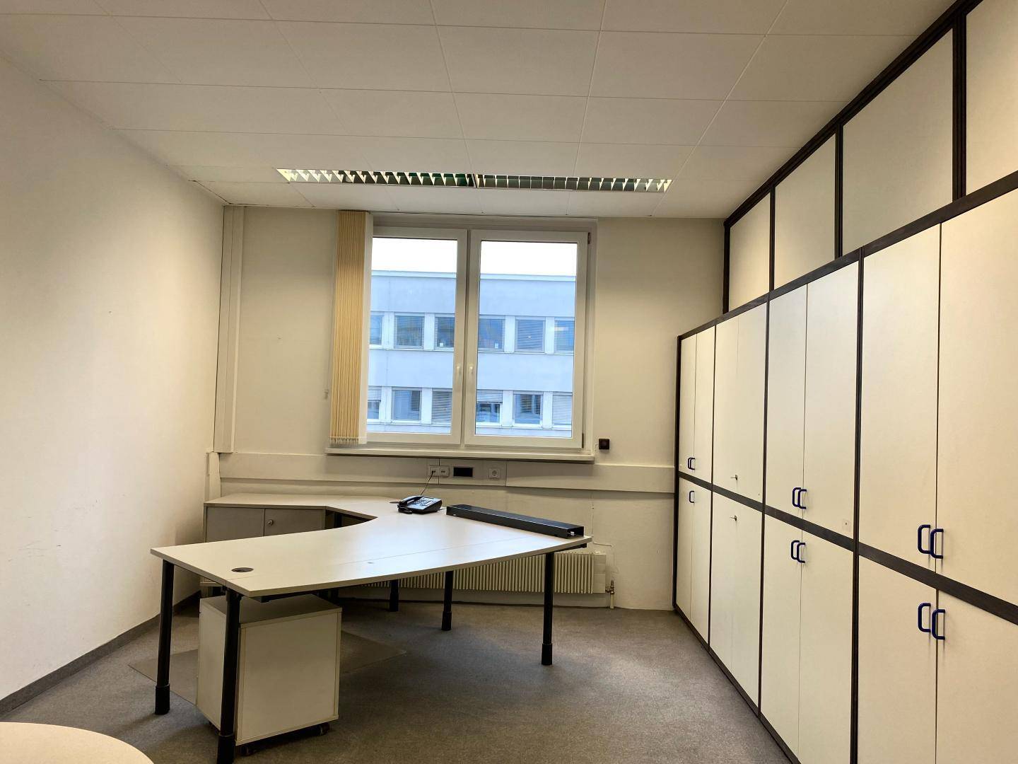 Büro mit einem großen Schreibtisch, integrierten Schränken und einem Fenster mit Blick auf ein Gebäude.