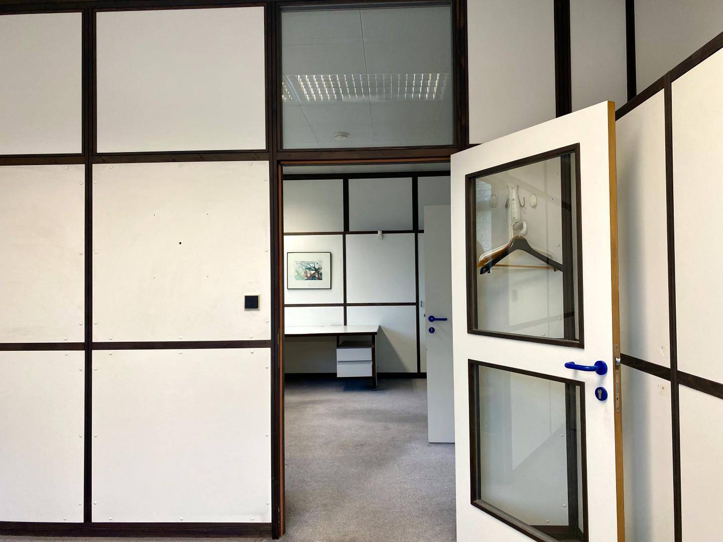 Blick durch eine offene Tür in ein weiteres Büro mit Schreibtisch und modularen Trennwänden.
