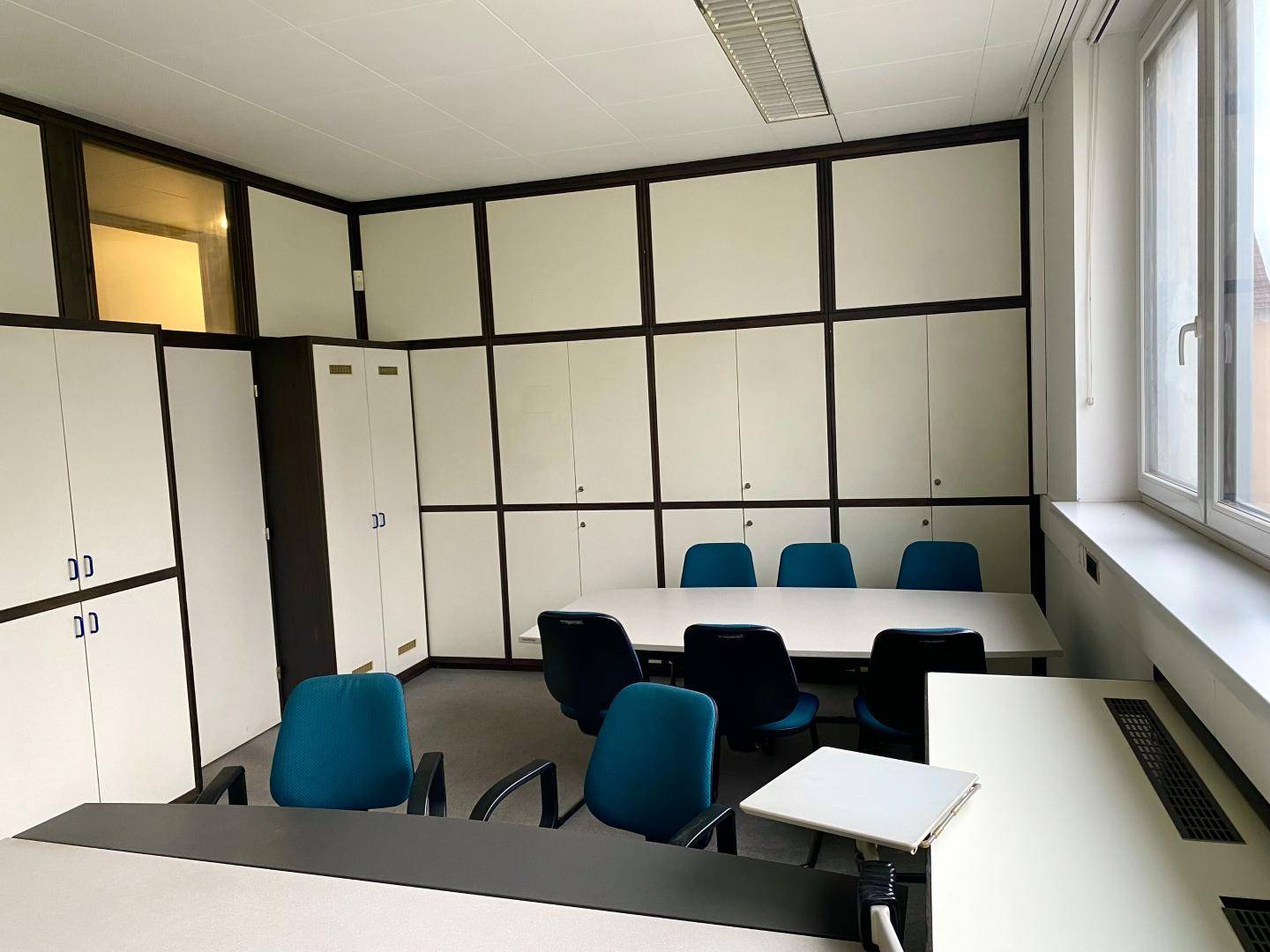 Konferenzraum mit einem langen Tisch, Stühlen und integrierten Schränken, mit Blick auf ein Gebäude.