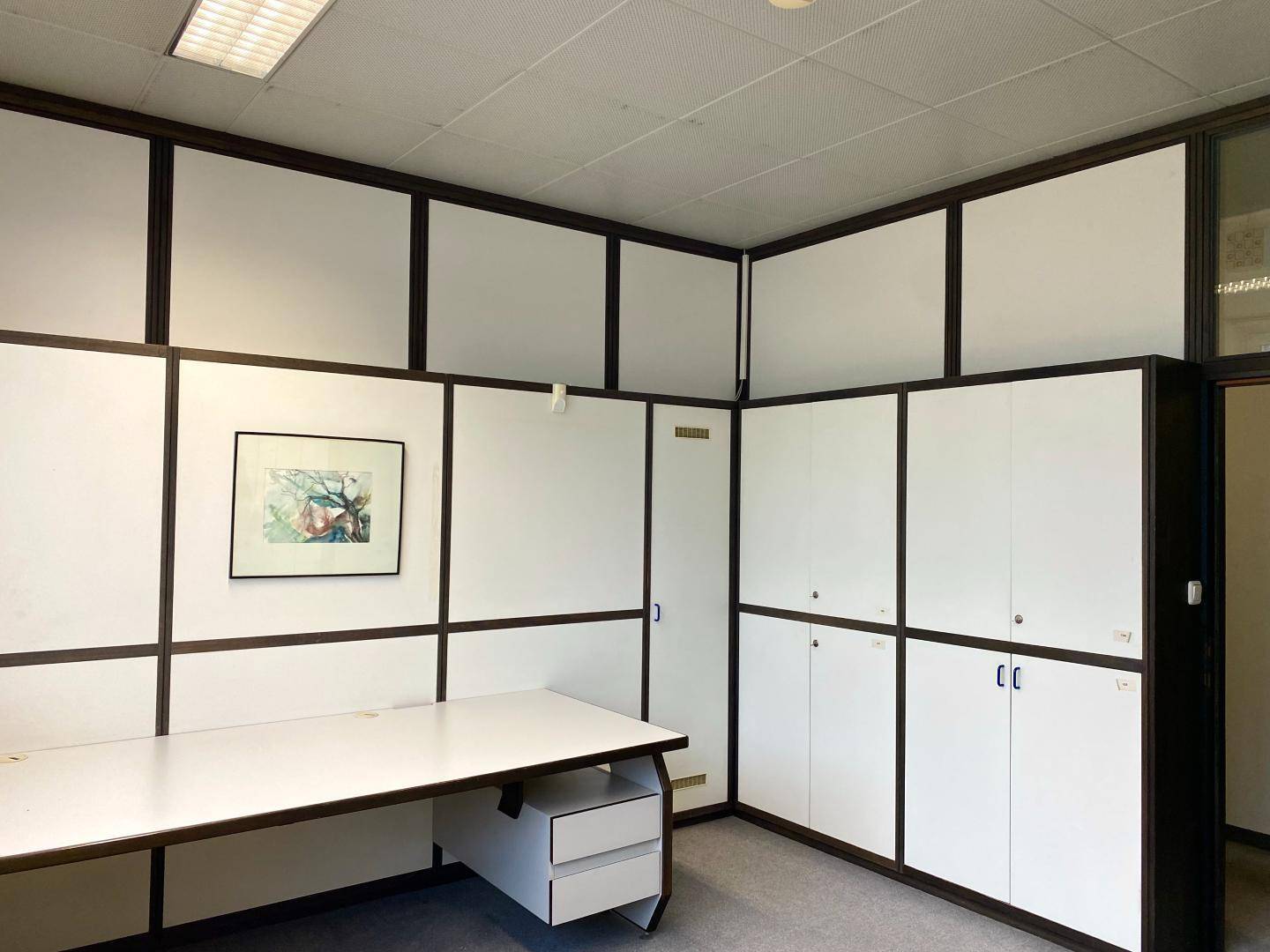 Büro mit einem Schreibtisch, integrierten Schränken und einem gerahmten Bild an der Wand.