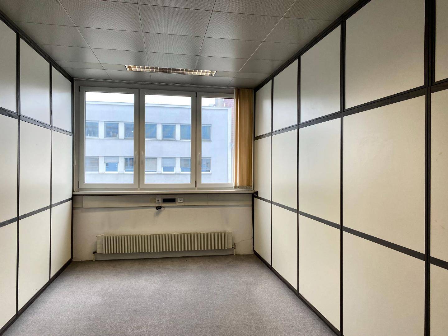 Leeres Büro mit modularen Trennwänden, Teppichboden und einem großen Fenster mit Blick nach draußen.