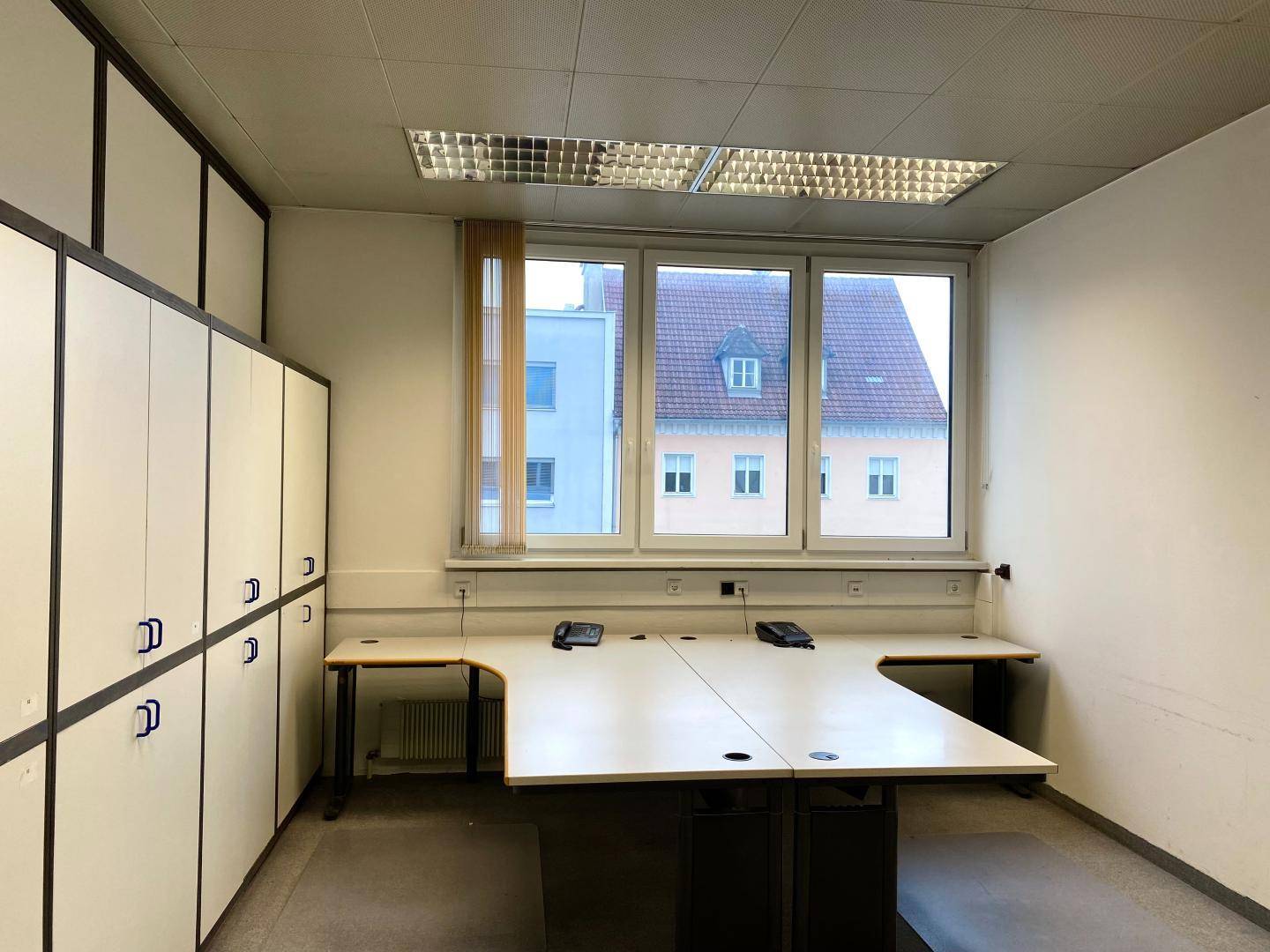 Büro mit zwei großen Schreibtischen, integrierten Schränken und einem Fenster mit Blick auf Dächer.