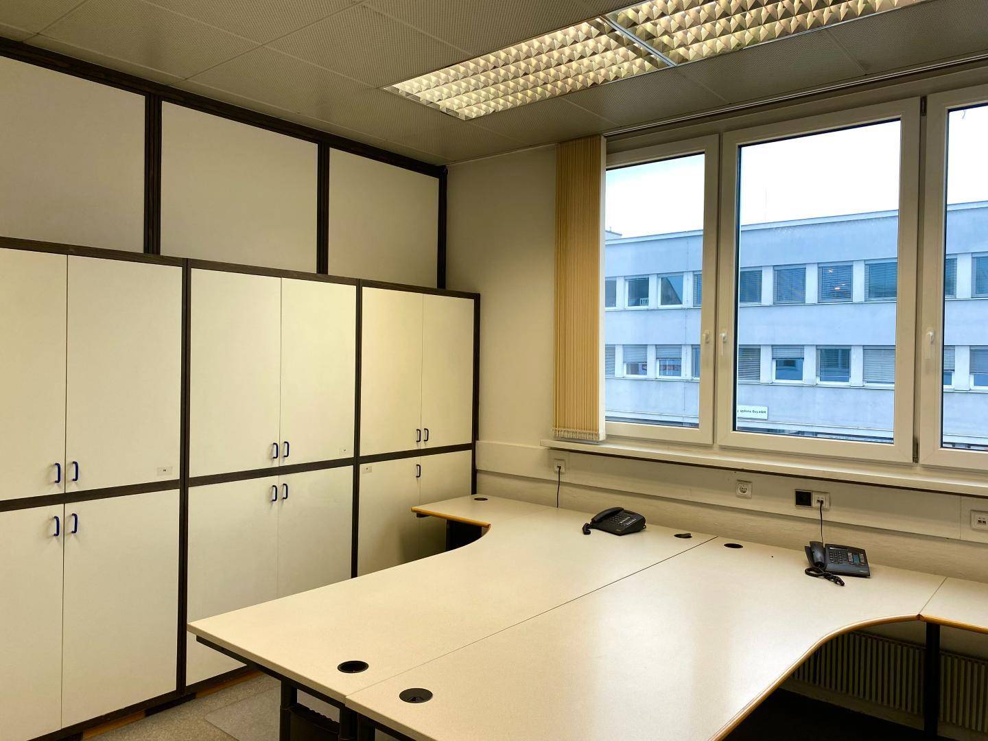 Büro mit zwei großen Schreibtischen, integrierten Schränken und einem Fenster mit Blick auf ein Gebäude.