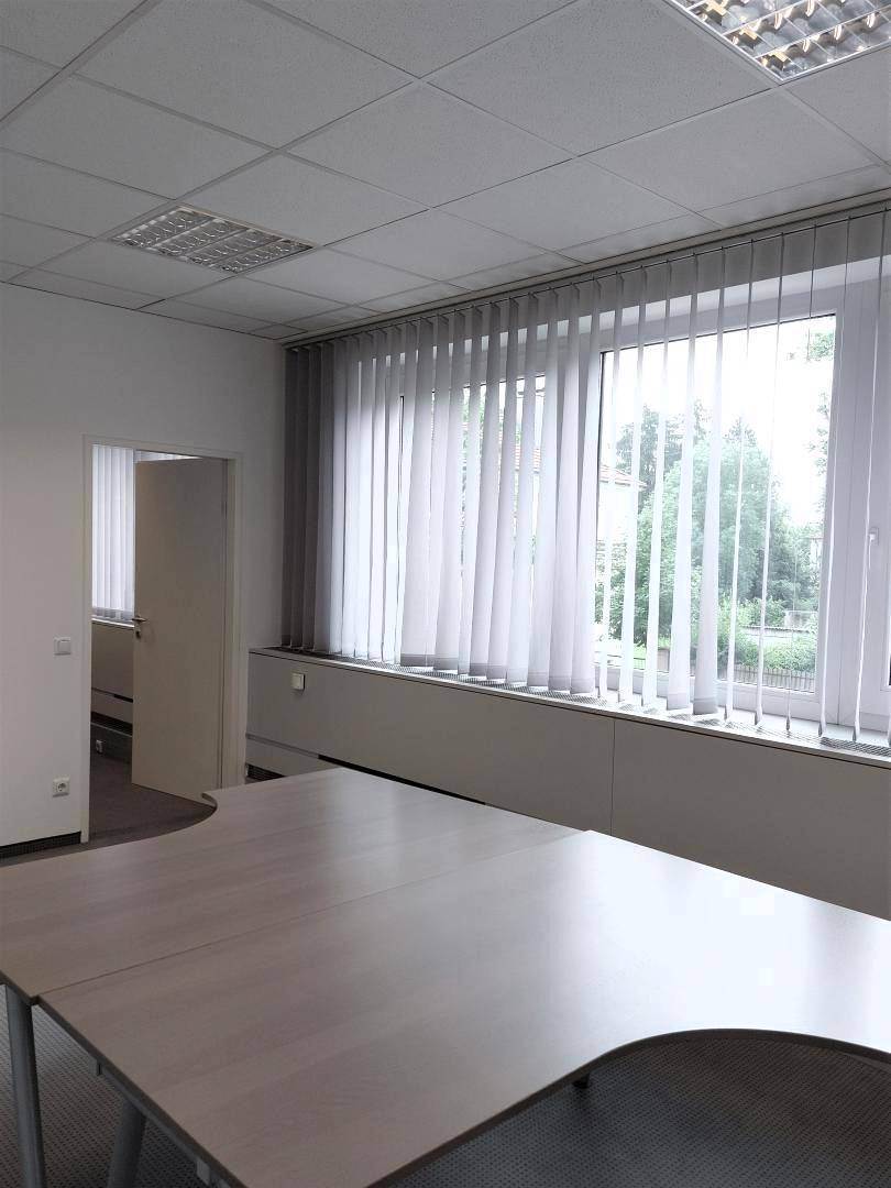 Heller Büroraum mit großen Fenstern und vertikalen Jalousien, die viel Tageslicht hereinlassen und eine grüne Aussicht bieten.