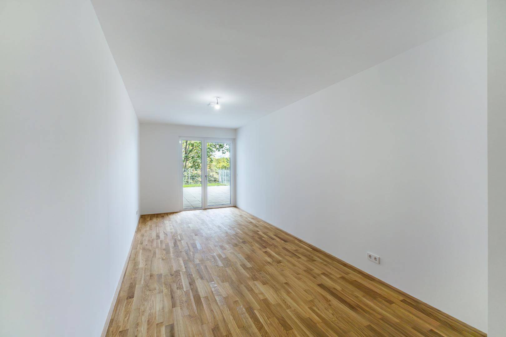 Heller, leerer Raum mit Holzboden und großem Fenster mit Zugang zu einem Außenbereich.