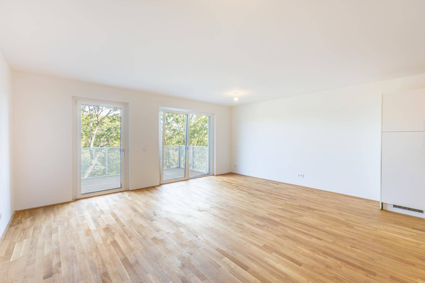 Heller Wohnraum mit Holzboden und zwei großen Glastüren, die zu einem Balkon führen.