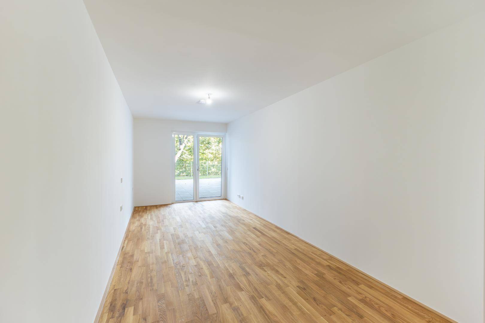 Leerer Raum mit hellem Parkettboden und Zugang zu einem Balkon oder einer Terrasse.