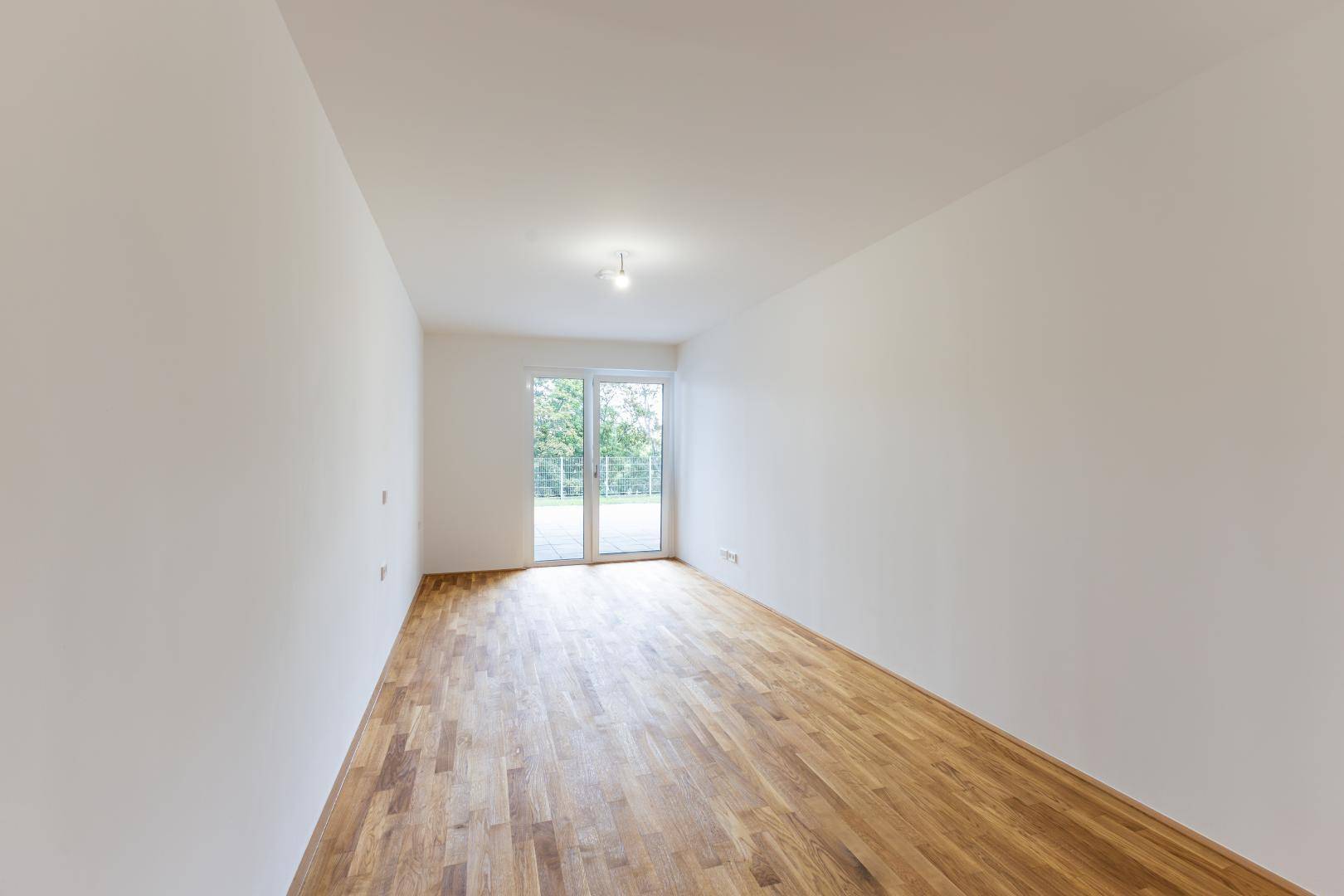 Heller, leerer Raum mit Holzboden und großen Fenstern, die Zugang zu einem Außenbereich bieten.