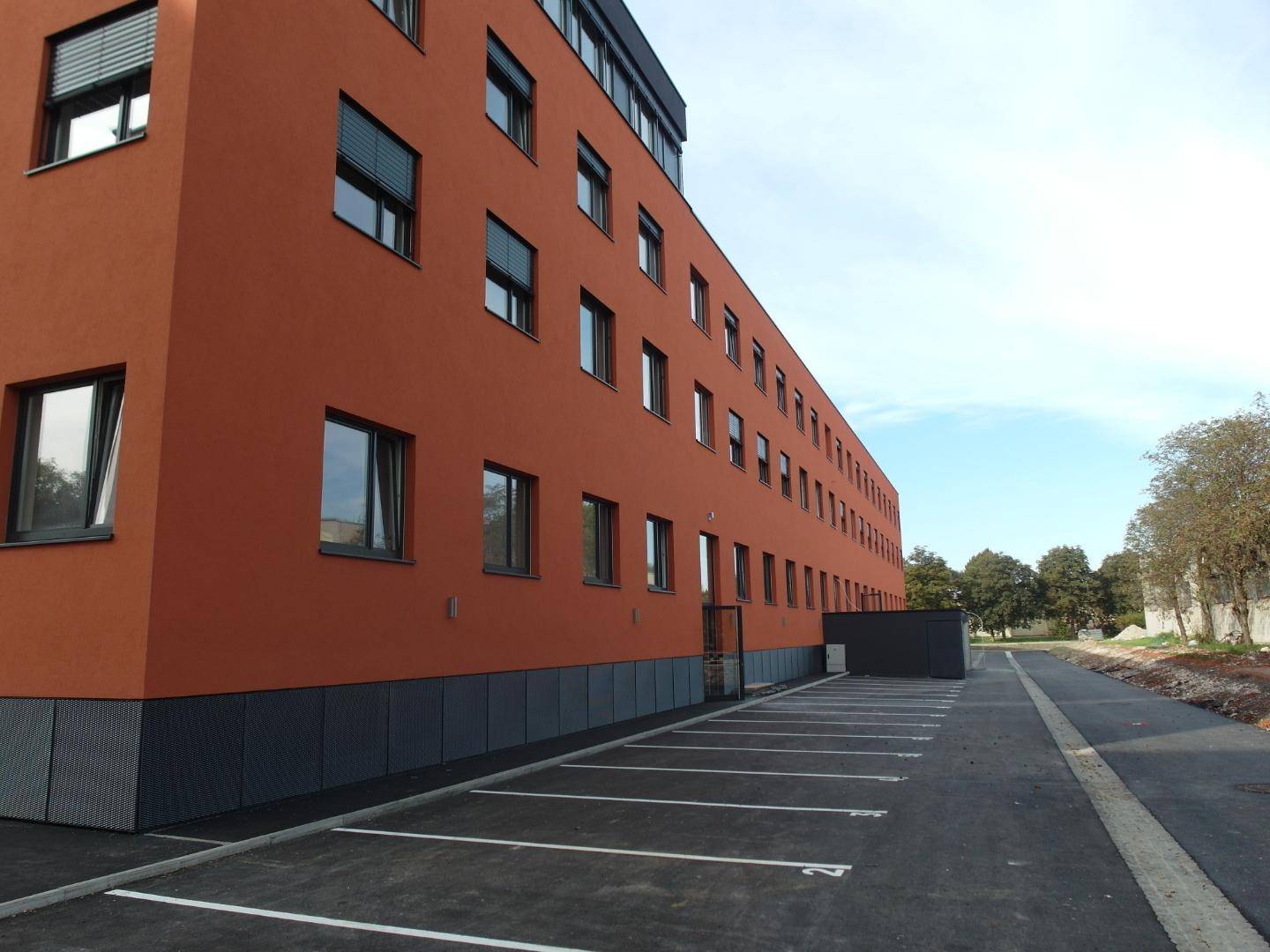 Seitenansicht eines modernen Gebäudes mit oranger Fassade und neu angelegten Parkplätzen.