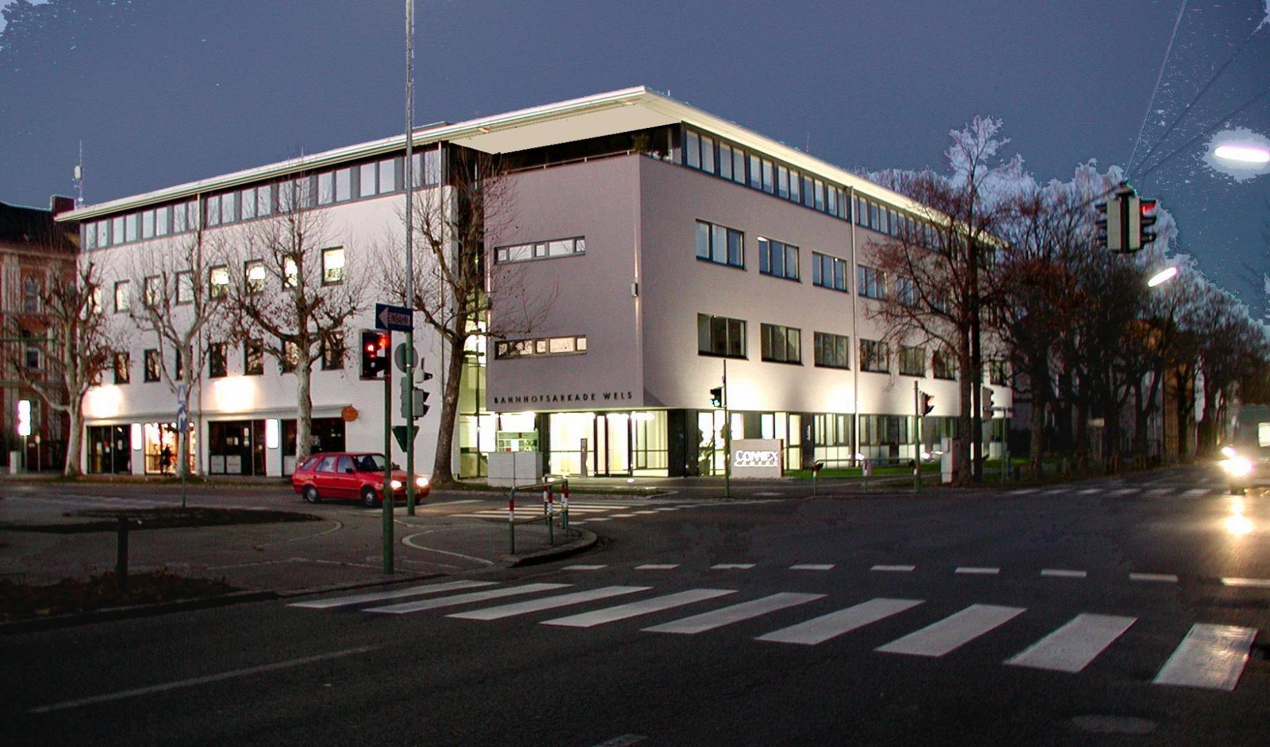 Die Bahnhofsarkade Wels bei Abendbeleuchtung, eingebettet in eine städtische Straßenszene.