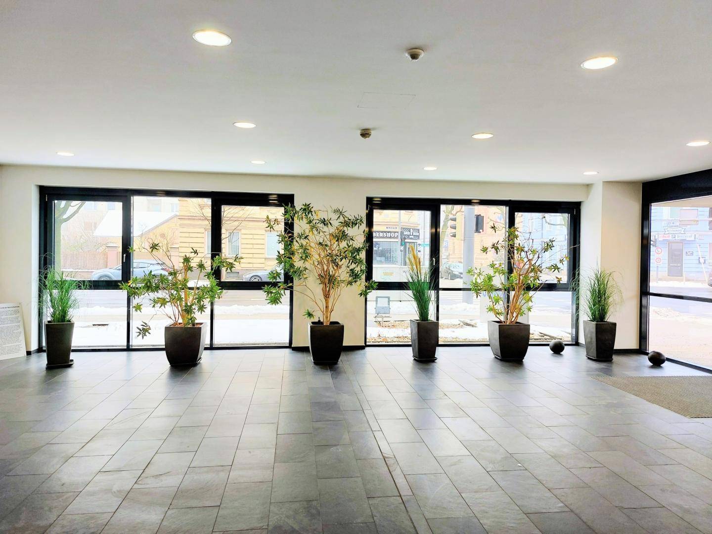 Heller Eingangsbereich mit großen Glasfronten, dunklen Fliesen und dekorativen Pflanzen, einladend gestaltet.
