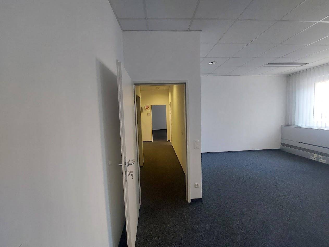 Blick aus einem Büroraum in einen Flur, beide Bereiche mit Teppichboden und weißen Wänden.