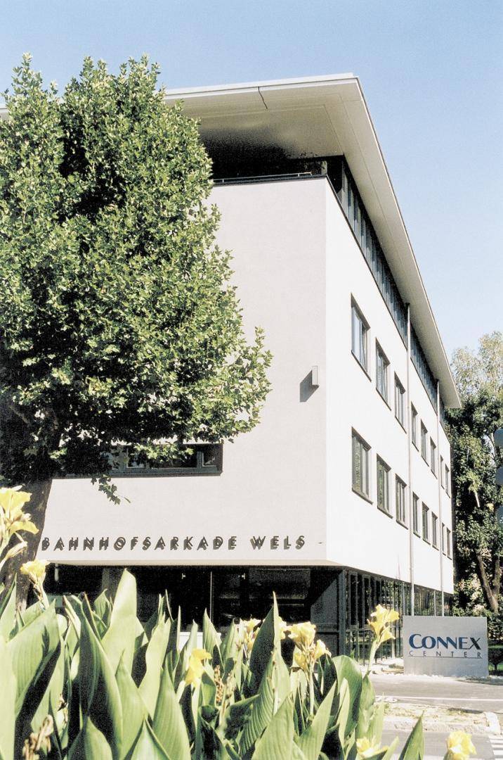 Moderne Geschäftsfassade der Bahnhofsarkade Wels mit Bäumen und Pflanzen im Vordergrund.
