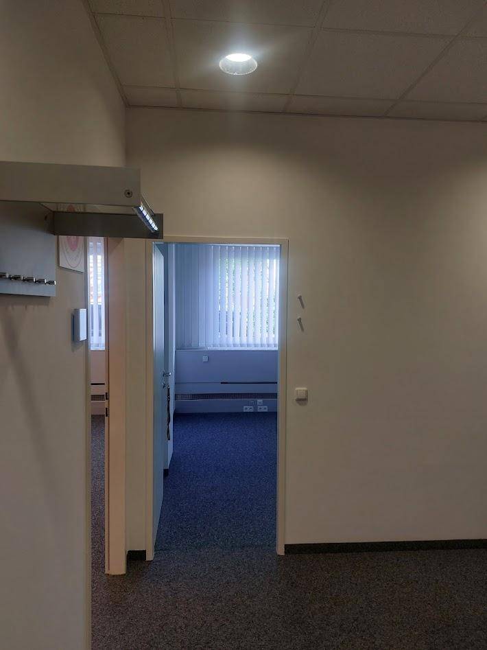 Blick von einem Teppichbereich durch eine offene Tür in einen weiteren Büroraum mit Fenstern.