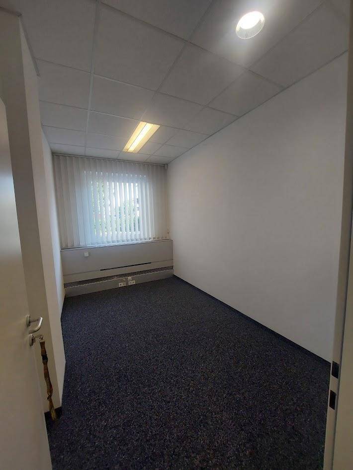 Kleinerer Büroraum mit Teppichboden, weißen Wänden und einem Fenster mit Jalousien.