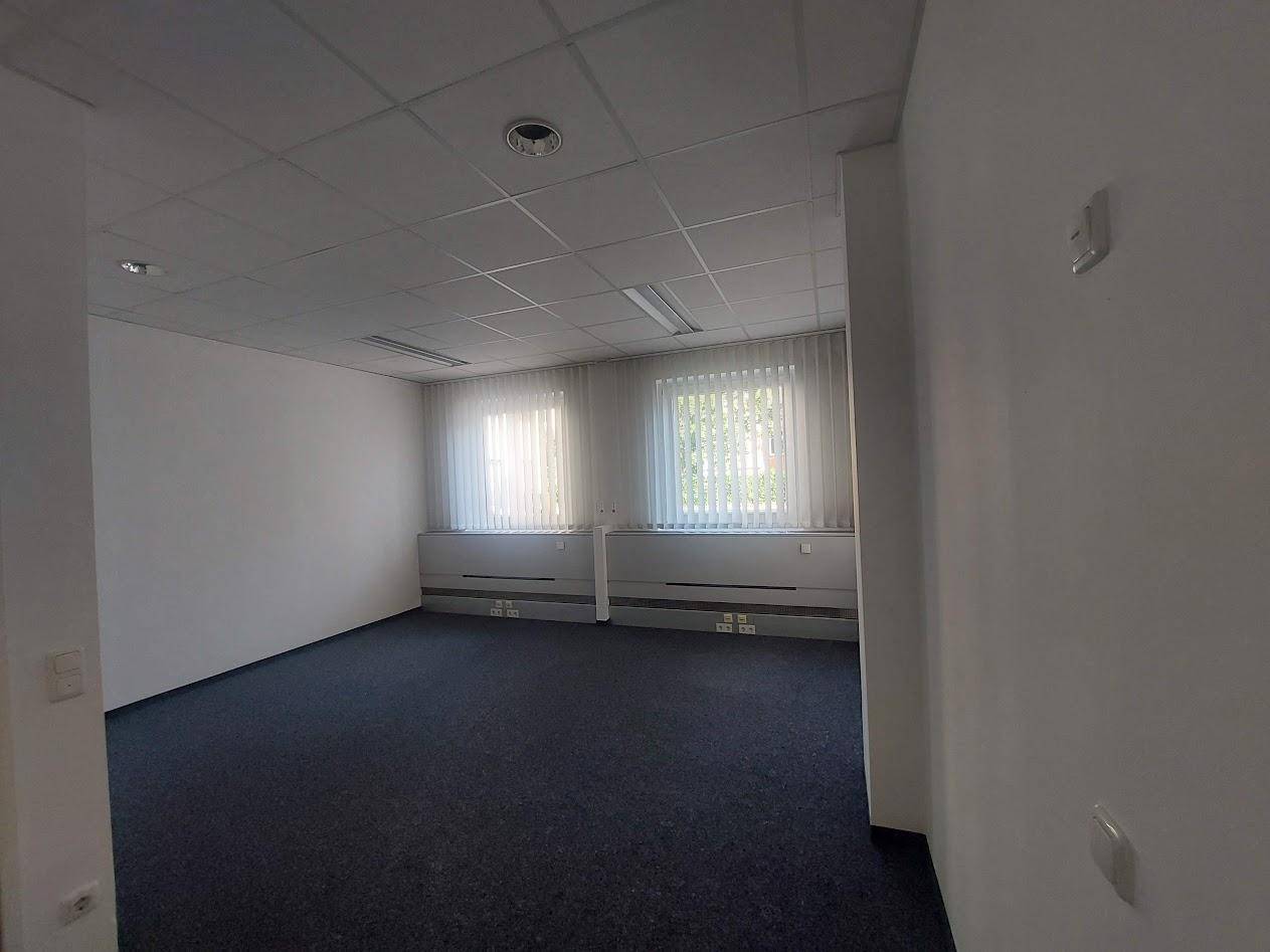 Heller Büroraum mit Teppichboden, weißen Wänden und zwei Fenstern, die viel Tageslicht hereinlassen.