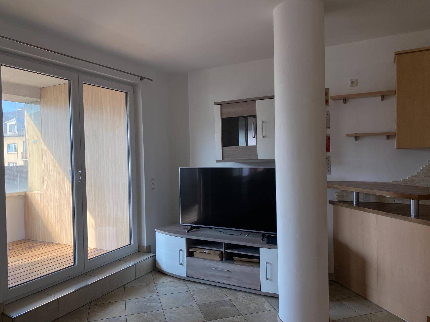 Wohnbereich mit Fernseher, einer Säule und Blick auf den Balkon, ausgestattet mit Fliesenboden.