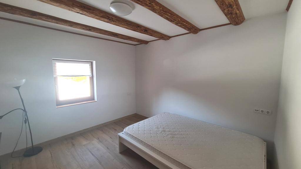 Schlafzimmer mit Holzbalken, Holzboden und einem Bett, hell durch das Fenster.