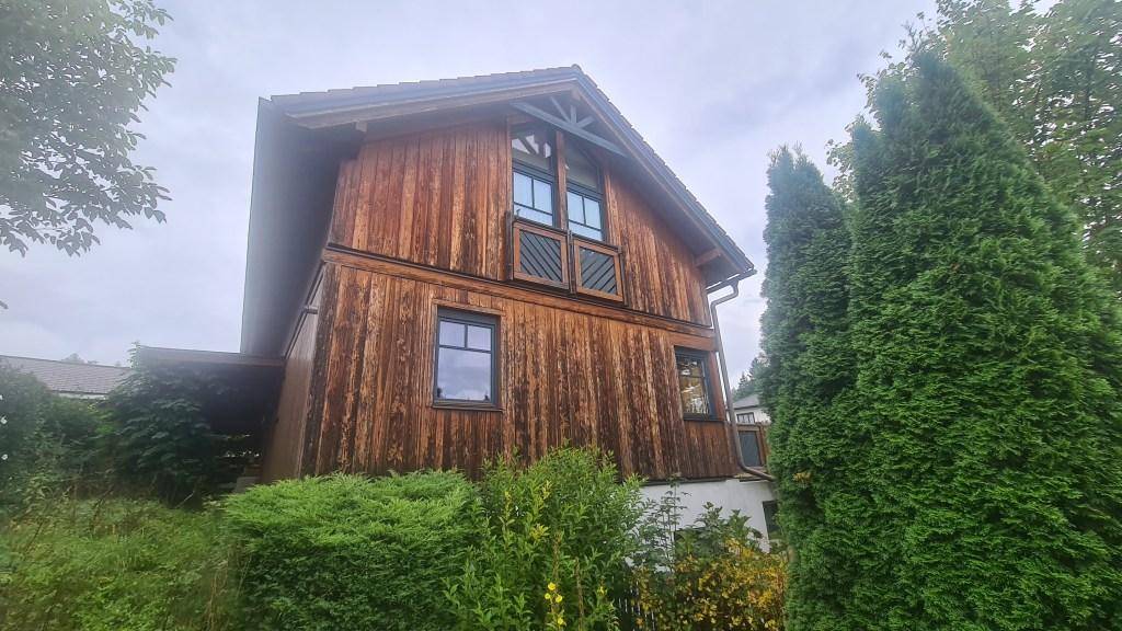 Seitenansicht eines Holzhauses mit hohen Nadelbäumen und traditioneller Holzfassade.