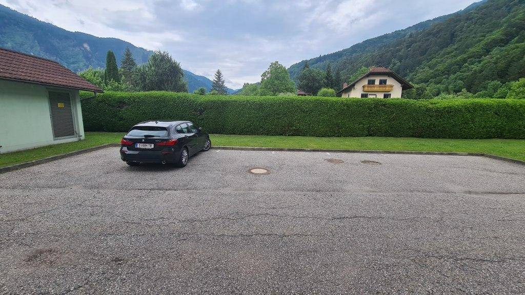 Offener Parkplatzbereich mit einem geparkten Auto, umgeben von Hecken und Bergen im Hintergrund.
