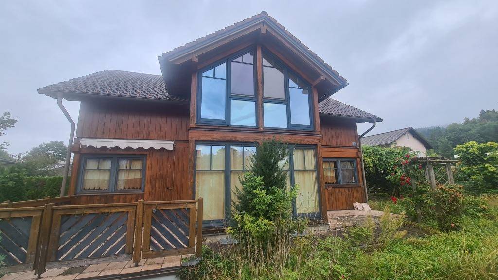 Holzhaus mit großen Giebelfenstern und einer kleinen Terrasse im Vordergrund, umgeben von Natur.