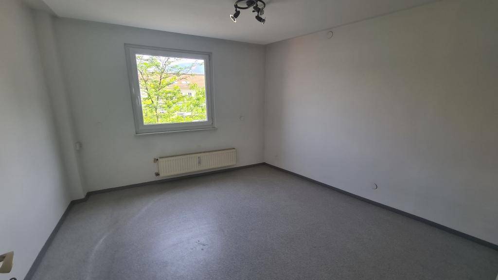 Schlichter Büroraum mit grauem Bodenbelag, weißen Wänden und einem Fenster mit Blick auf Bäume.