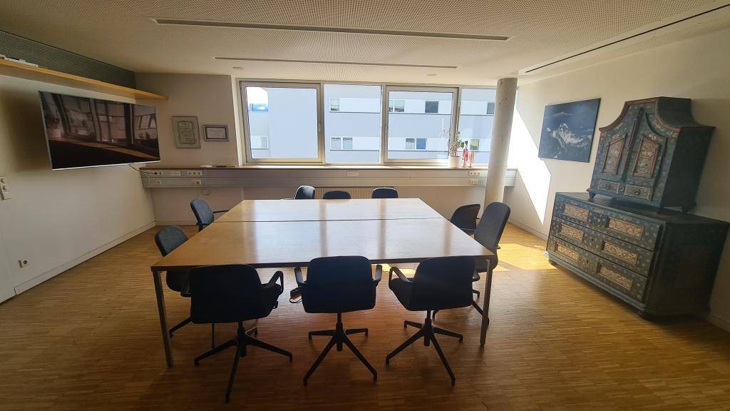 Konferenzraum mit großem Holztisch, bequemen Stühlen und einem dekorativen Schrank, beleuchtet durch breite Fenster.