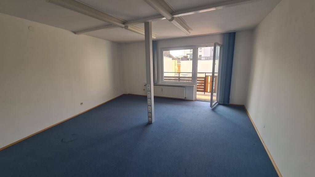 Büroraum mit blauem Teppichboden, weißen Wänden und einer Säule, mit direktem Zugang zum Balkon.