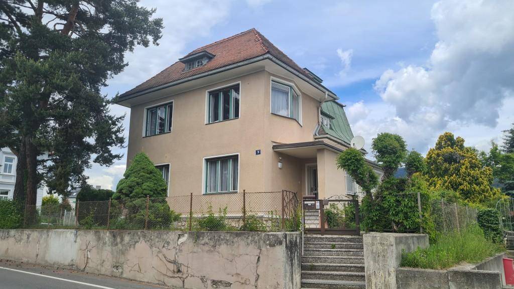 Vorderansicht des Hauses mit beigefarbener Fassade, Satteldach und altem Baumbestand unter bewölktem Himmel.