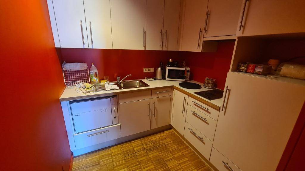 Kleine Küchenzeile mit weißen Schränken, roter Wand und integrierten Geräten, ideal für eine schnelle Mahlzeit im Büro.