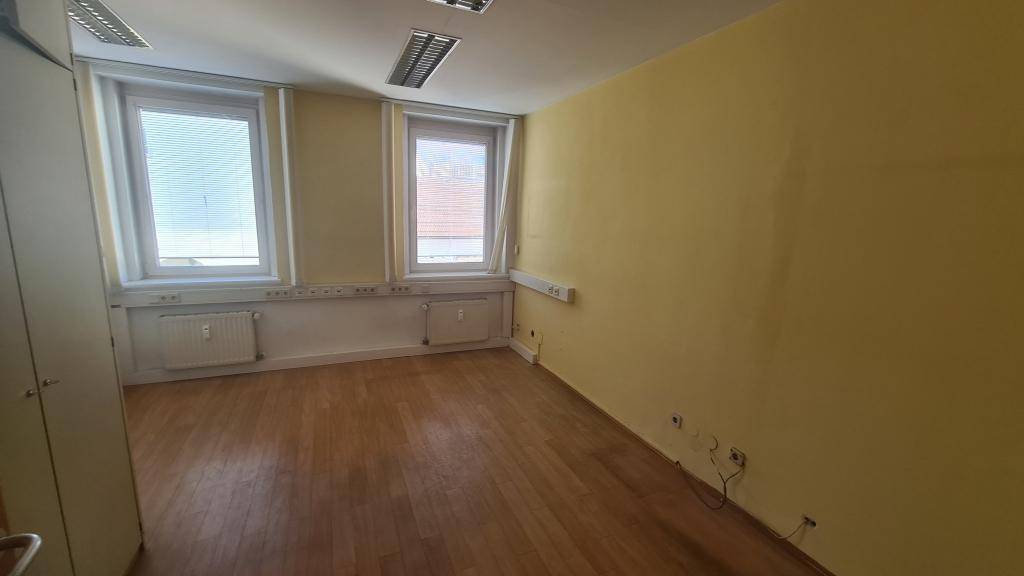 Helles Büro mit Holzboden, zwei großen Fenstern und gelber Akzentwand, ideal für kreative Arbeitsbereiche.