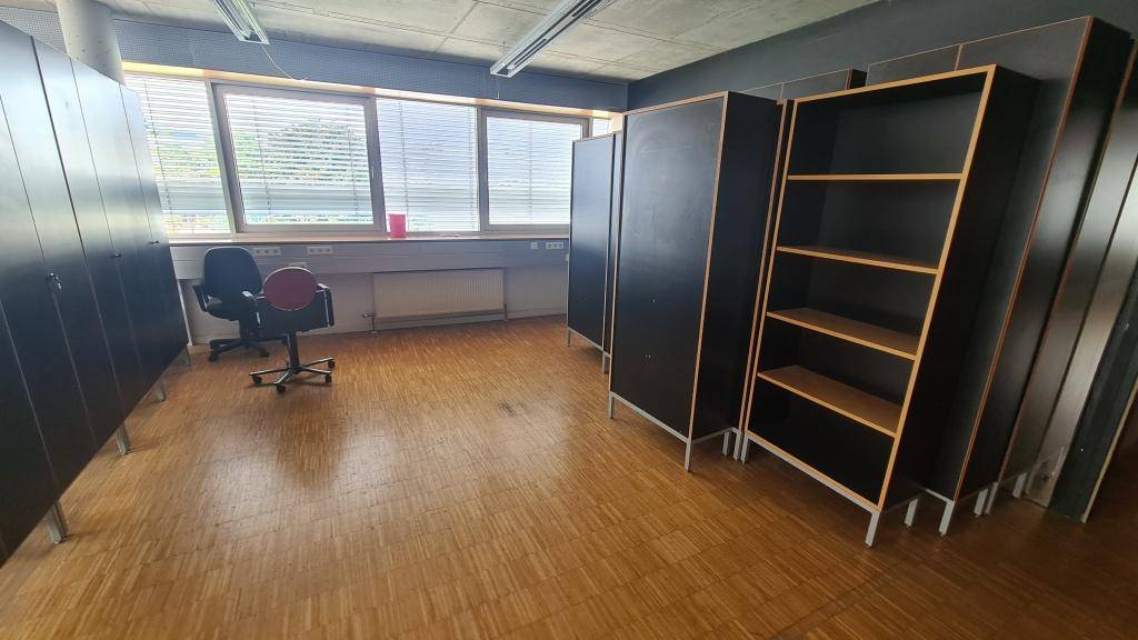 Offener Bürobereich mit Holzparkettboden, großen Fenstern und schwarzen Regalen, die den Raum funktional unterteilen.