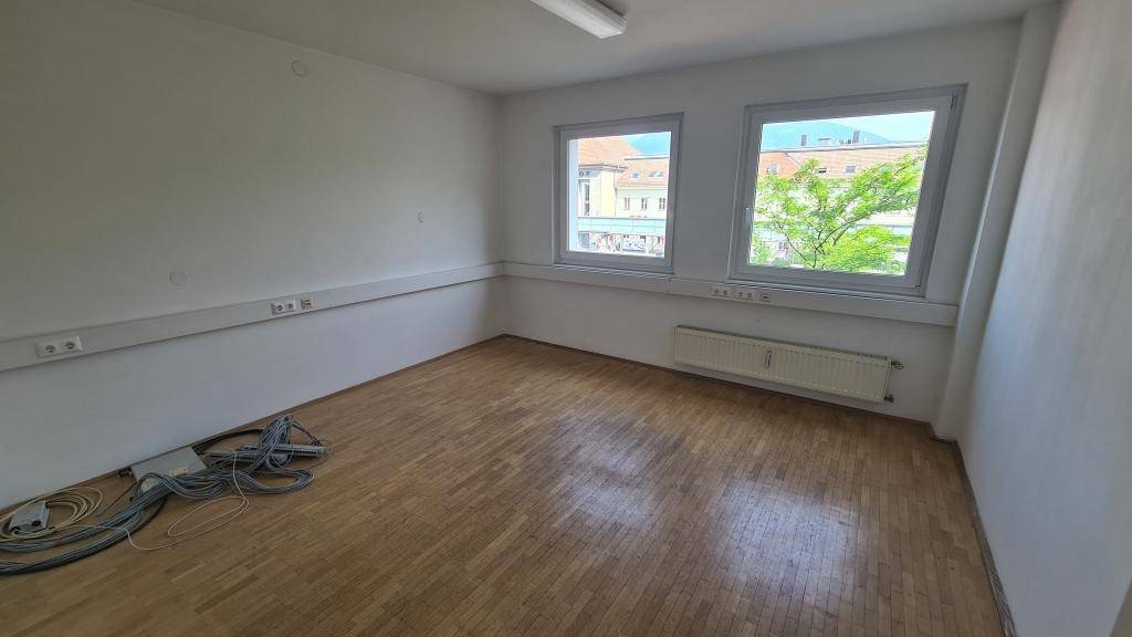 Leerer Büroraum mit Parkettboden, weißen Wänden und zwei Fenstern mit Blick auf die Stadt.