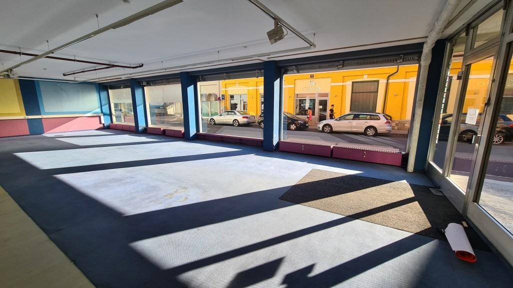 Großer Gewerberaum mit blauem Teppichboden und weitem Blick auf die Straße durch die Schaufenster.